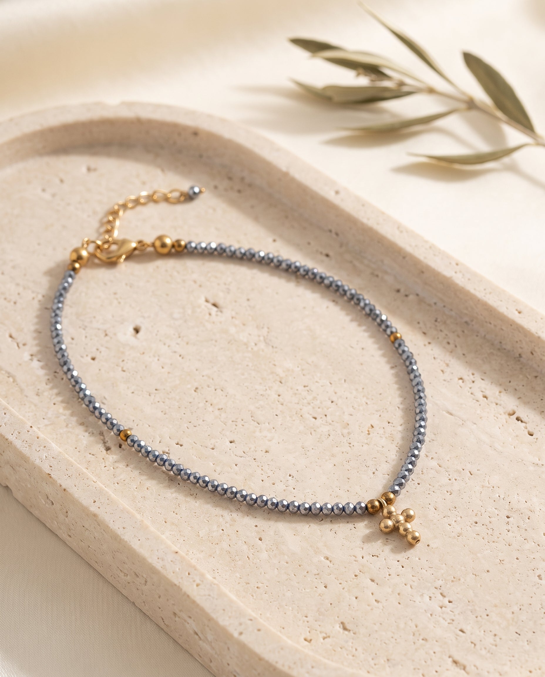 Collar cruz de cristal facetado azul acero sobre bandeja de piedra beige con rama de olivo, Perla Santa