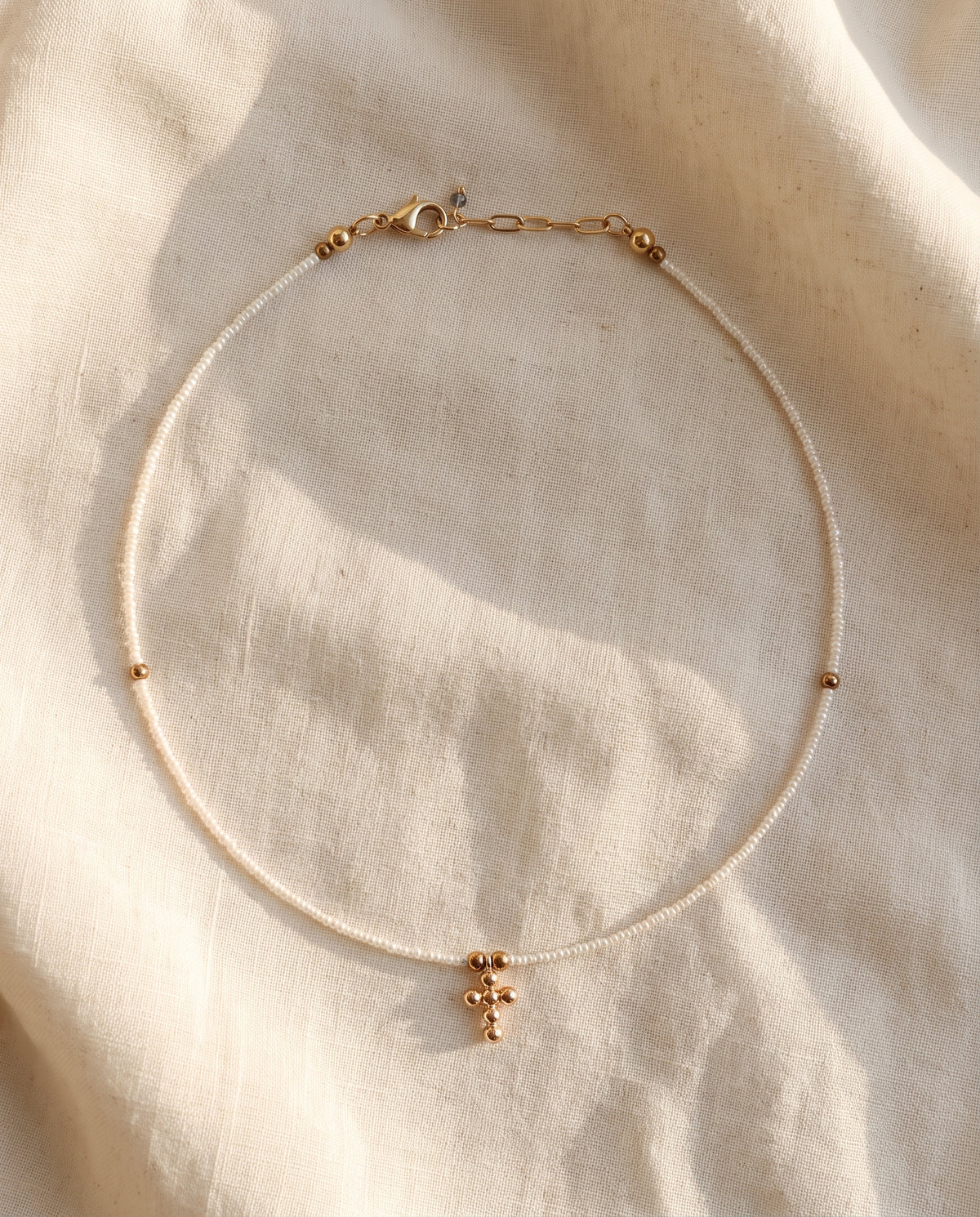 Collar cruz de perla natural blanca sobre lino crema con luz calida, Perla Santa