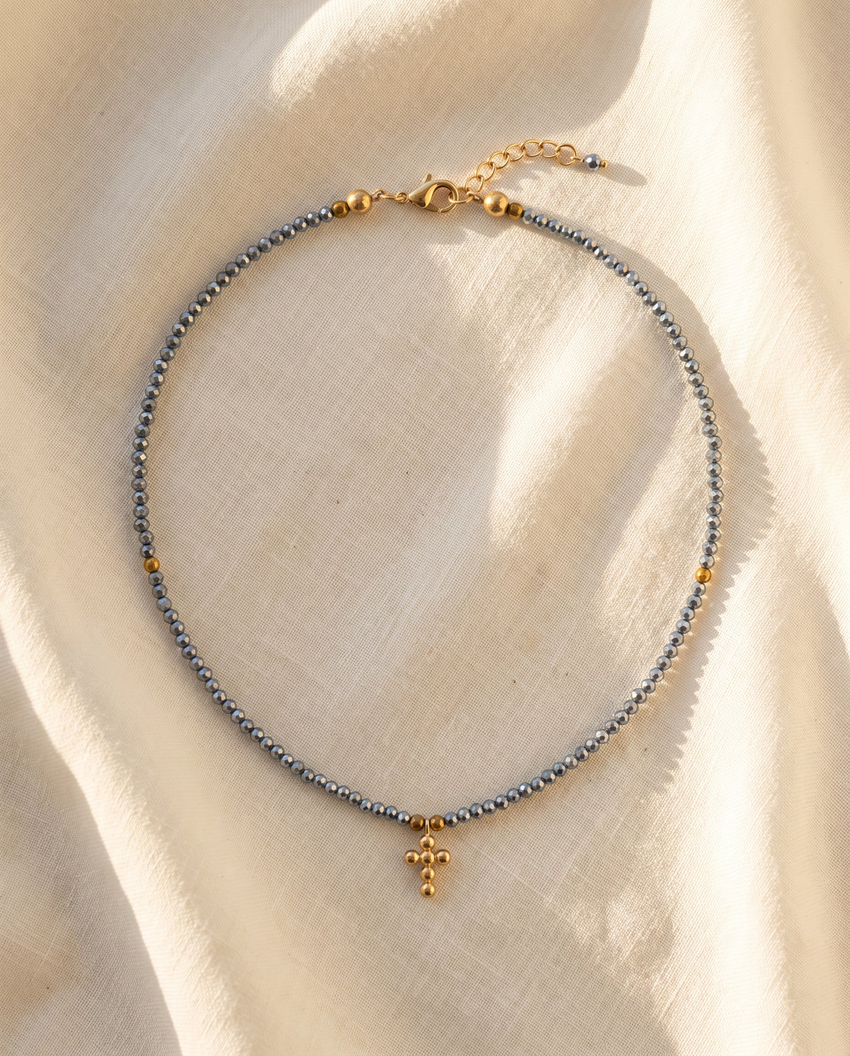 Collar cruz de cristal facetado azul acero sobre lino natural crema con luz calida, Perla Santa