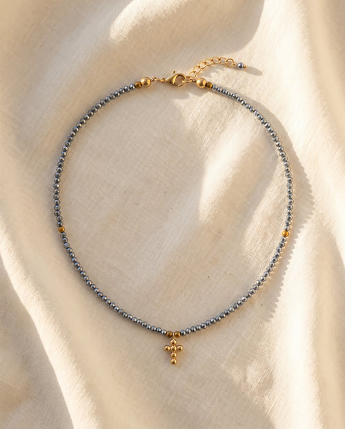 Collar cruz de cristal facetado azul acero sobre lino natural crema con luz calida, Perla Santa