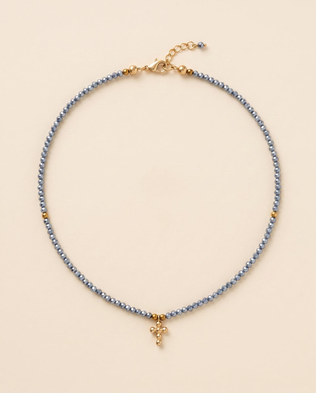 Collar de cristal facetado azul acero con cruz de chapa de oro sobre fondo crema, Perla Santa
