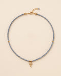 Collar de cristal facetado azul acero con cruz de chapa de oro sobre fondo crema, Perla Santa