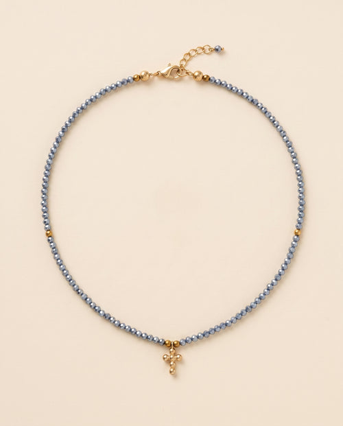 Collar de cristal facetado azul acero con cruz de chapa de oro sobre fondo crema, Perla Santa