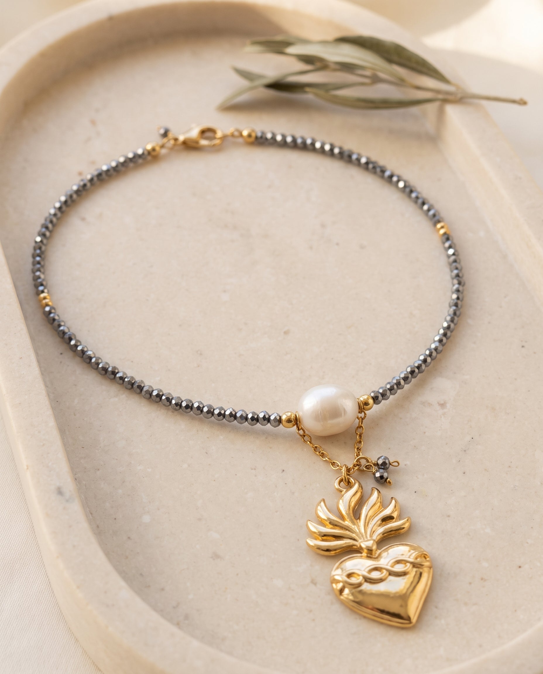 Collar de corazón Perla Santa sobre bandeja de piedra beige con rama de olivo