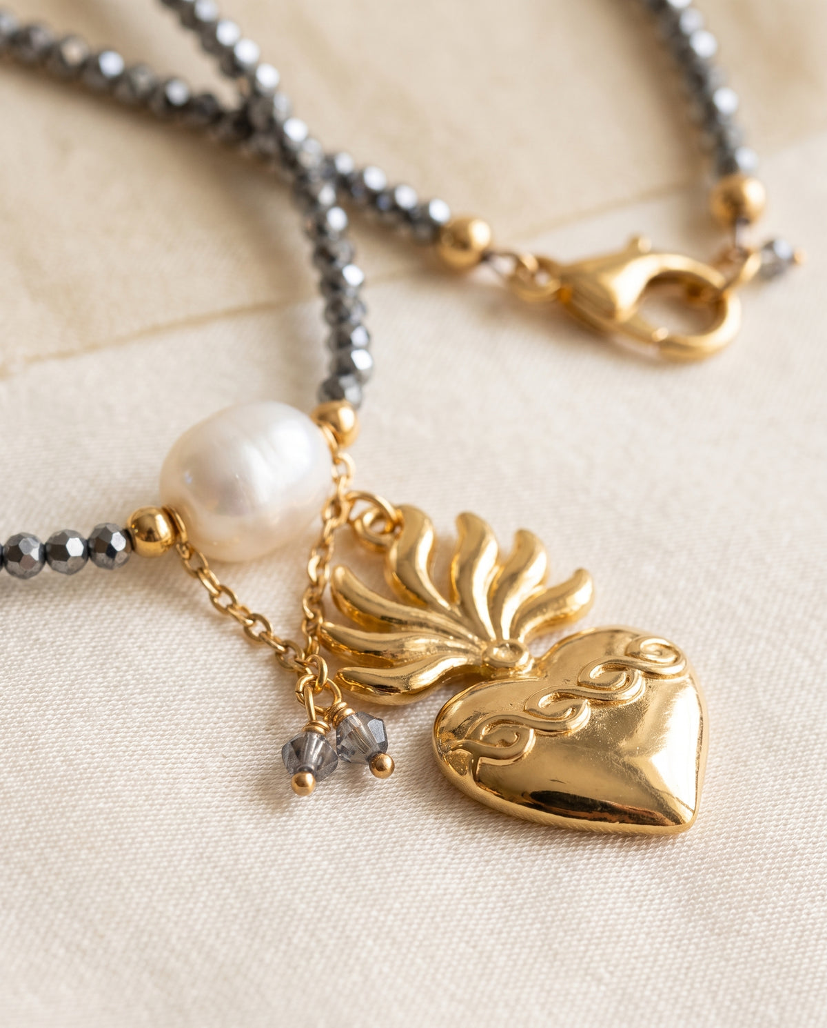 Detalle del dije de corazón chapado en oro con llamas y de la perla natural blanca del collar Perla Santa