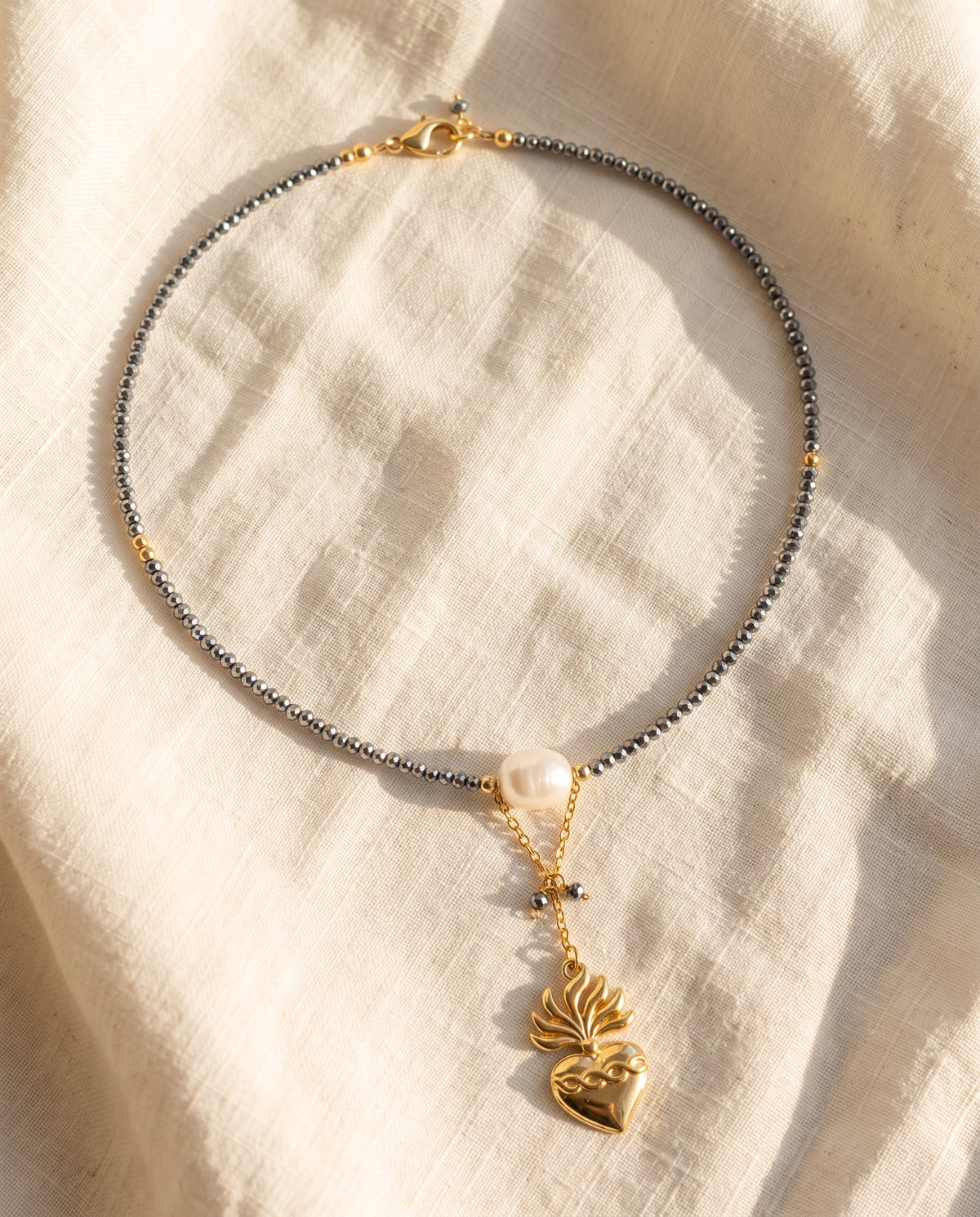 Collar de corazón chapado en oro y cristal facetado gris de Perla Santa sobre lino natural color crema
