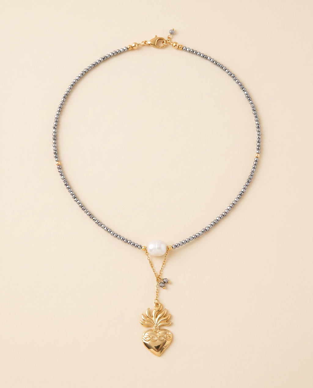 Collar Perla Santa con hilo de cristal facetado gris, perla natural blanca y dije de corazón chapado en oro, sobre fondo crema
