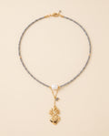 Collar Perla Santa con hilo de cristal facetado gris, perla natural blanca y dije de corazón chapado en oro, sobre fondo crema