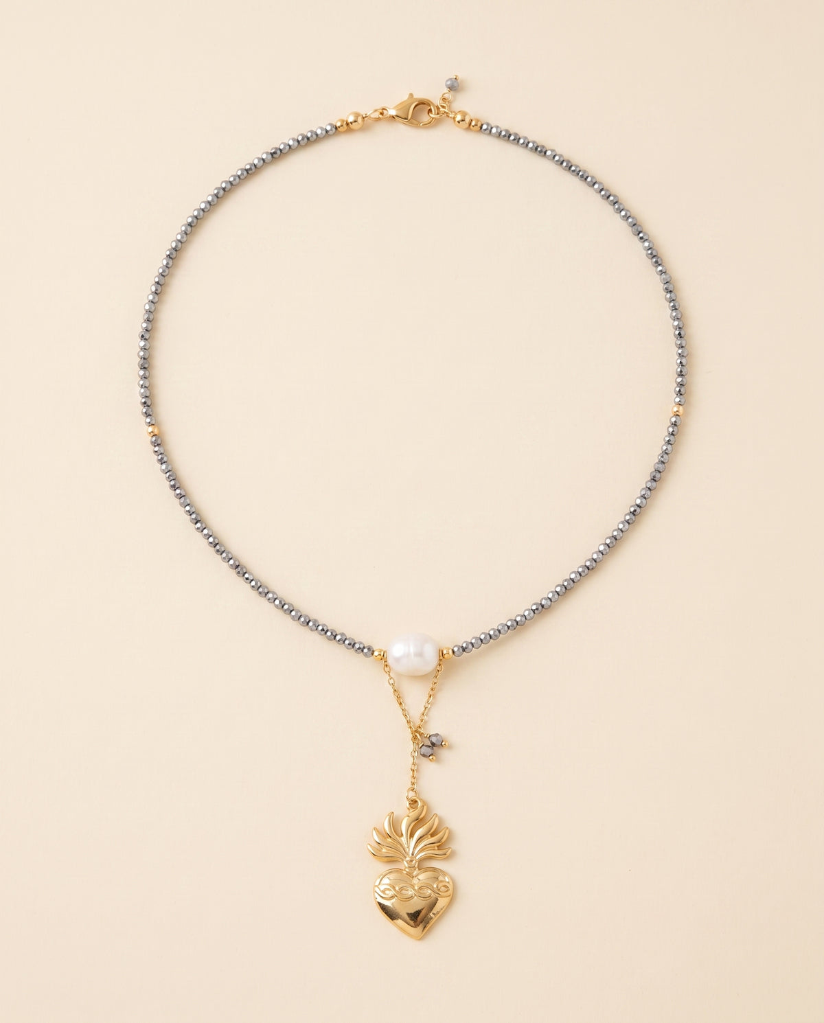 Collar Perla Santa con hilo de cristal facetado gris, perla natural blanca y dije de corazón chapado en oro, sobre fondo crema