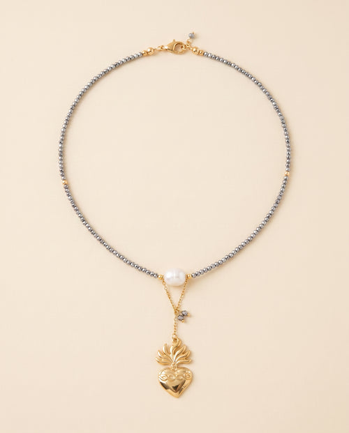 Collar Perla Santa con hilo de cristal facetado gris, perla natural blanca y dije de corazón chapado en oro, sobre fondo crema