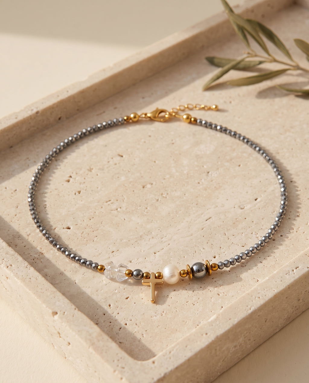 Collar de cristal con cruz dorada de Perla Santa sobre bandeja de piedra beige con rama de olivo