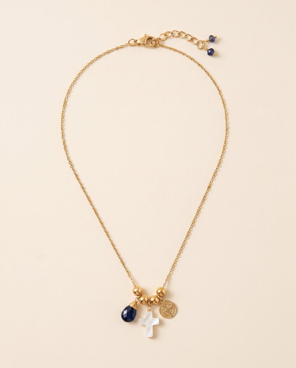 Collar Perla Santa de cadena dorada con cruz de concha nácar, medalla de San Benito y piedra azul facetada, sobre fondo crema