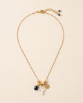 Collar Perla Santa de cadena dorada con cruz de concha nácar, medalla de San Benito y piedra azul facetada, sobre fondo crema