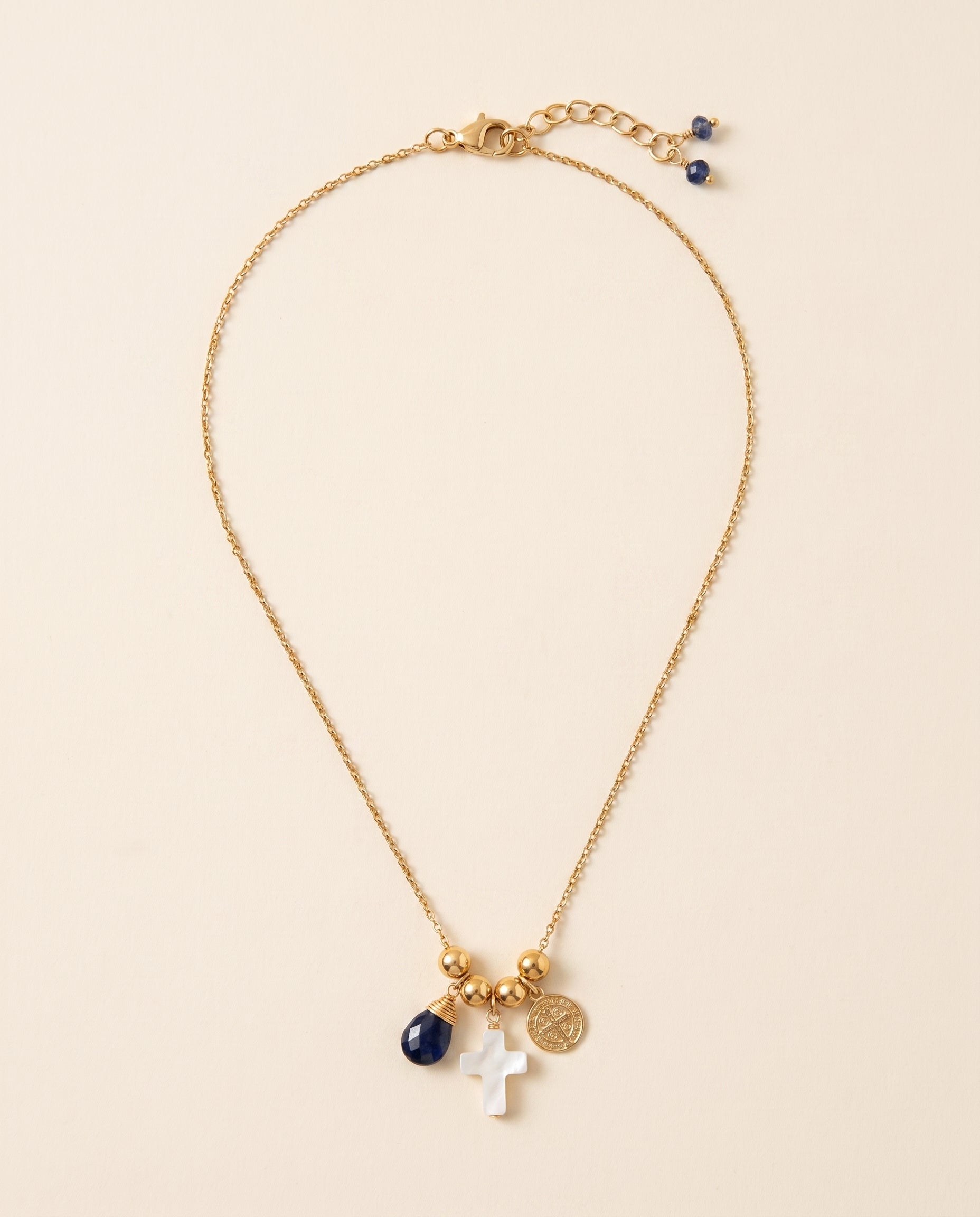 Collar Perla Santa de cadena dorada con cruz de concha nácar, medalla de San Benito y piedra azul facetada, sobre fondo crema