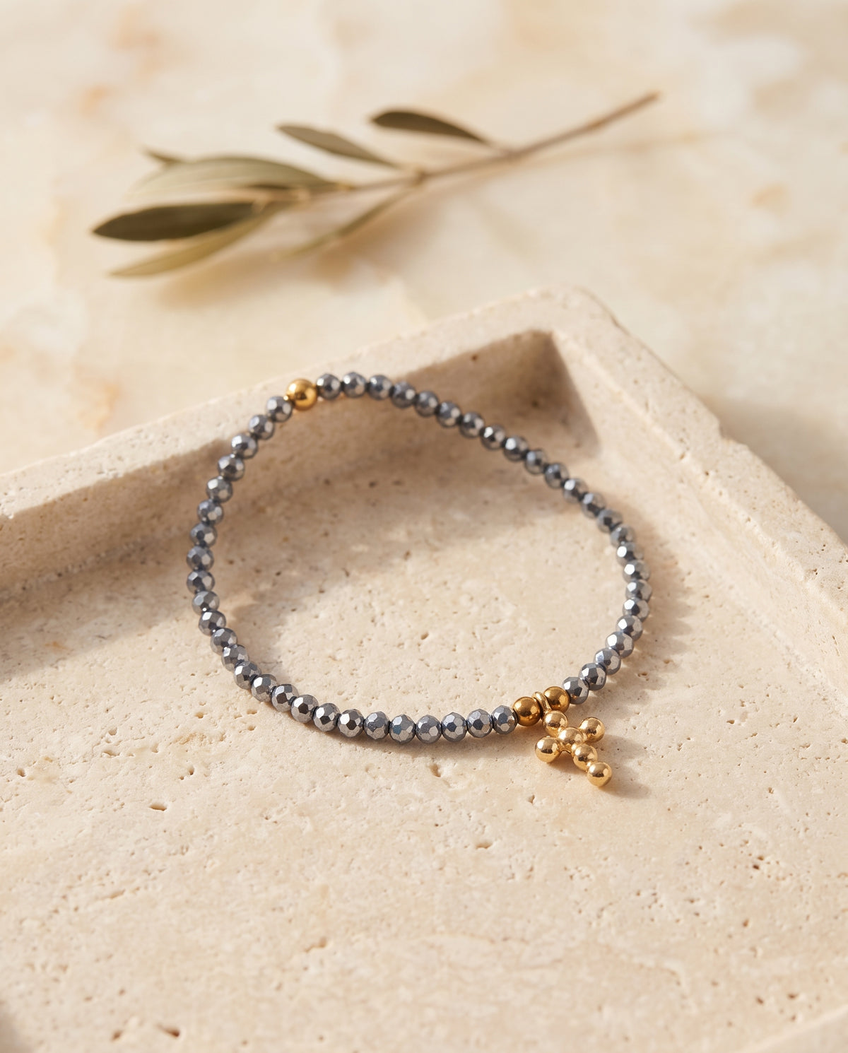 Pulsera cruz de cristal facetado azul acero sobre bandeja de piedra beige con rama de olivo, Perla Santa