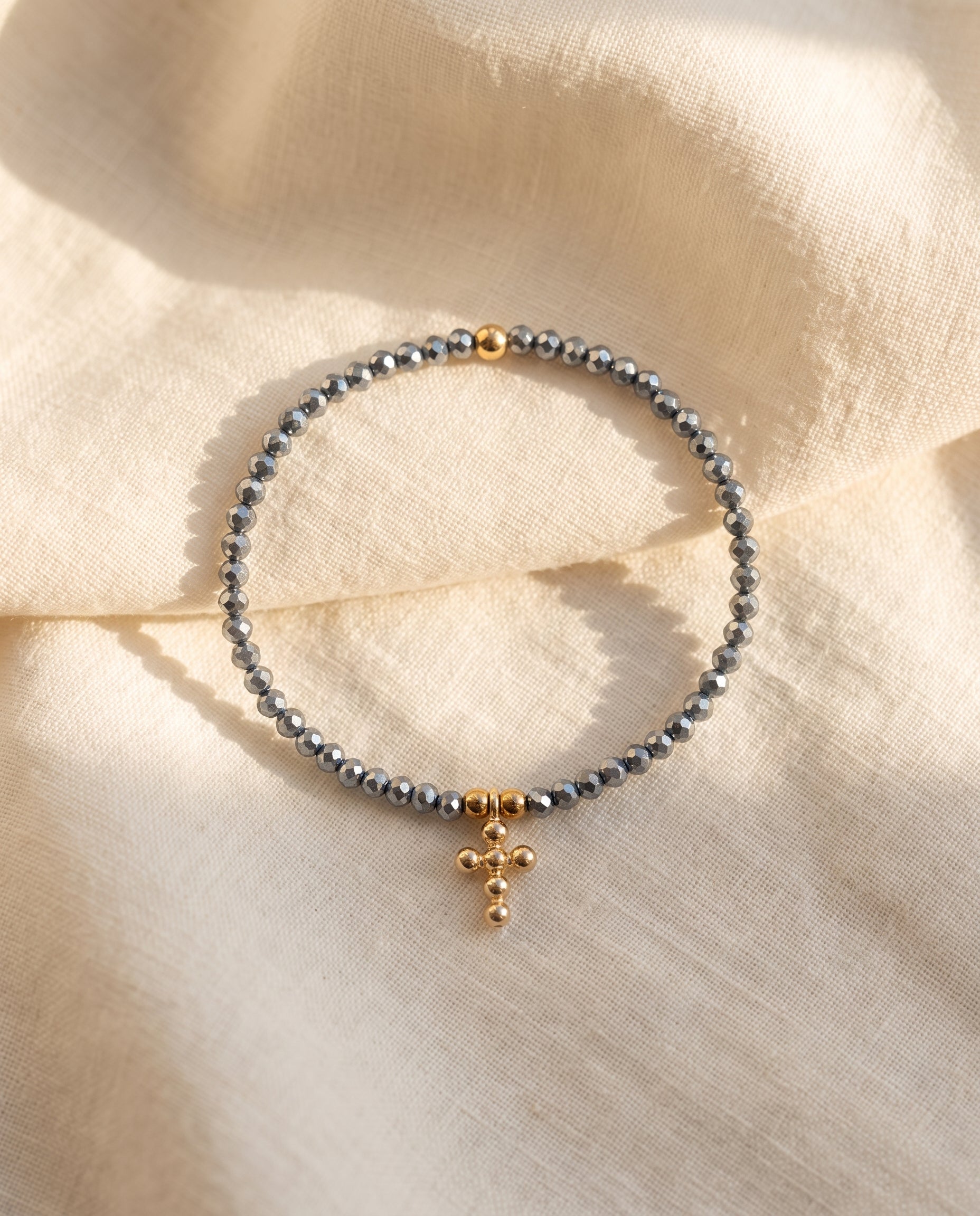 Pulsera cruz de cristal facetado azul acero sobre lino crema con luz calida, Perla Santa