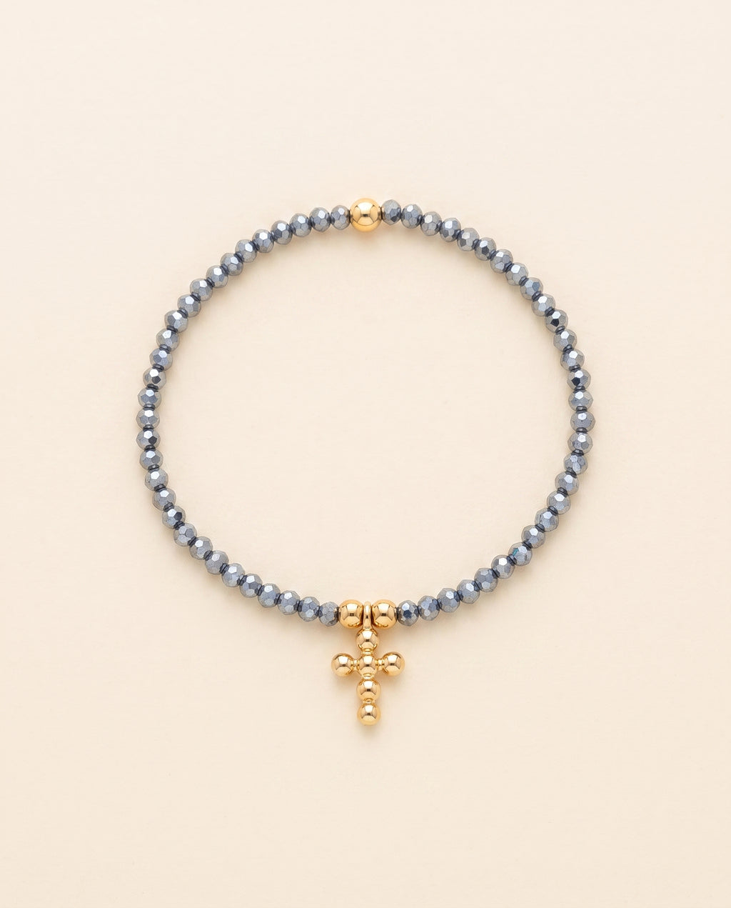 Pulsera de cristal facetado azul acero con cruz de chapa de oro sobre fondo crema, Perla Santa