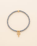 Pulsera de cristal facetado azul acero con cruz de chapa de oro sobre fondo crema, Perla Santa