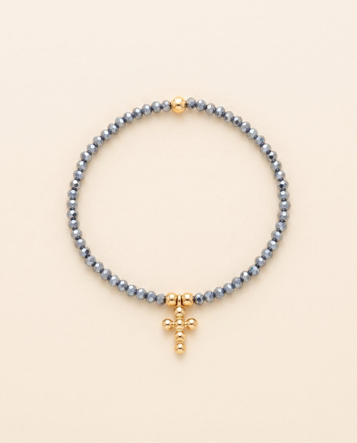 Pulsera de cristal facetado azul acero con cruz de chapa de oro sobre fondo crema, Perla Santa