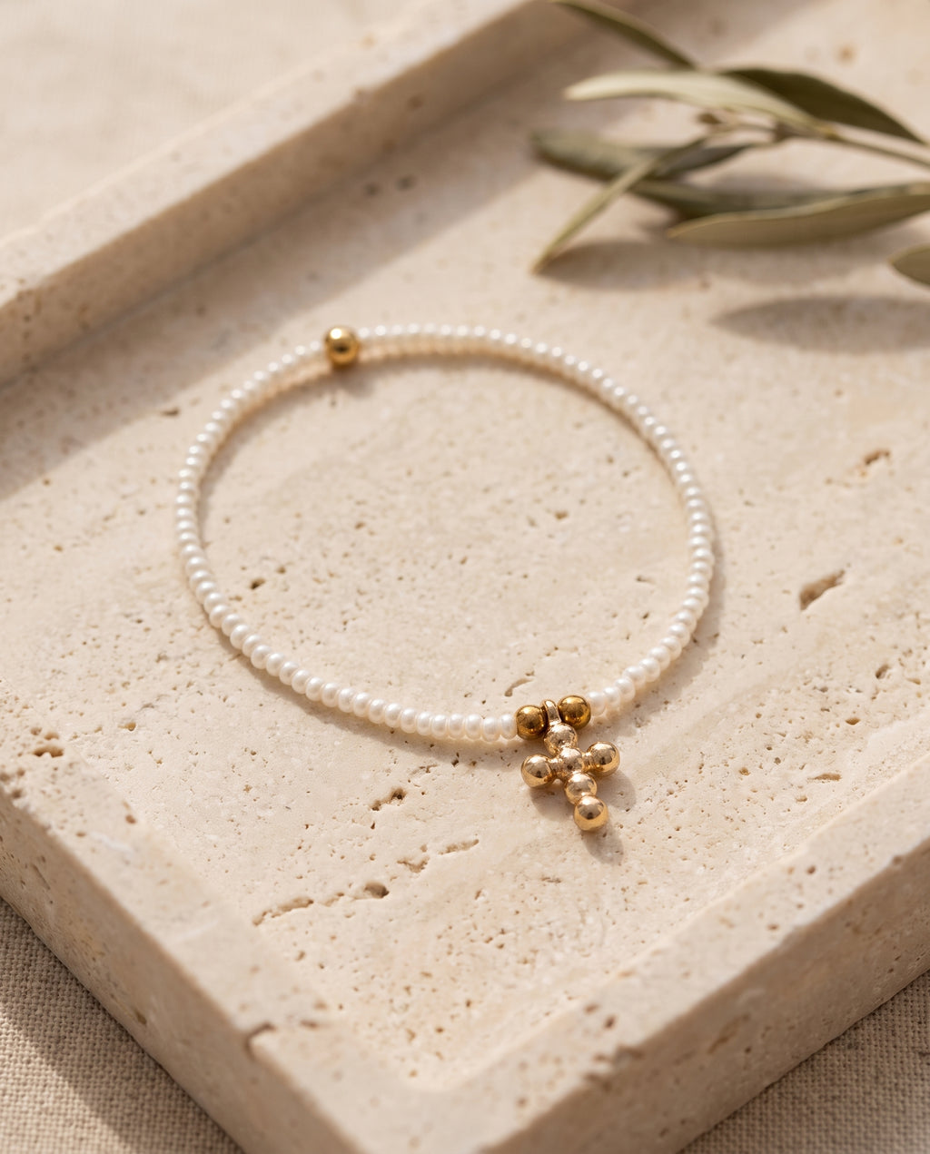 Pulsera cruz de perla natural blanca sobre bandeja de piedra beige con rama de olivo, Perla Santa