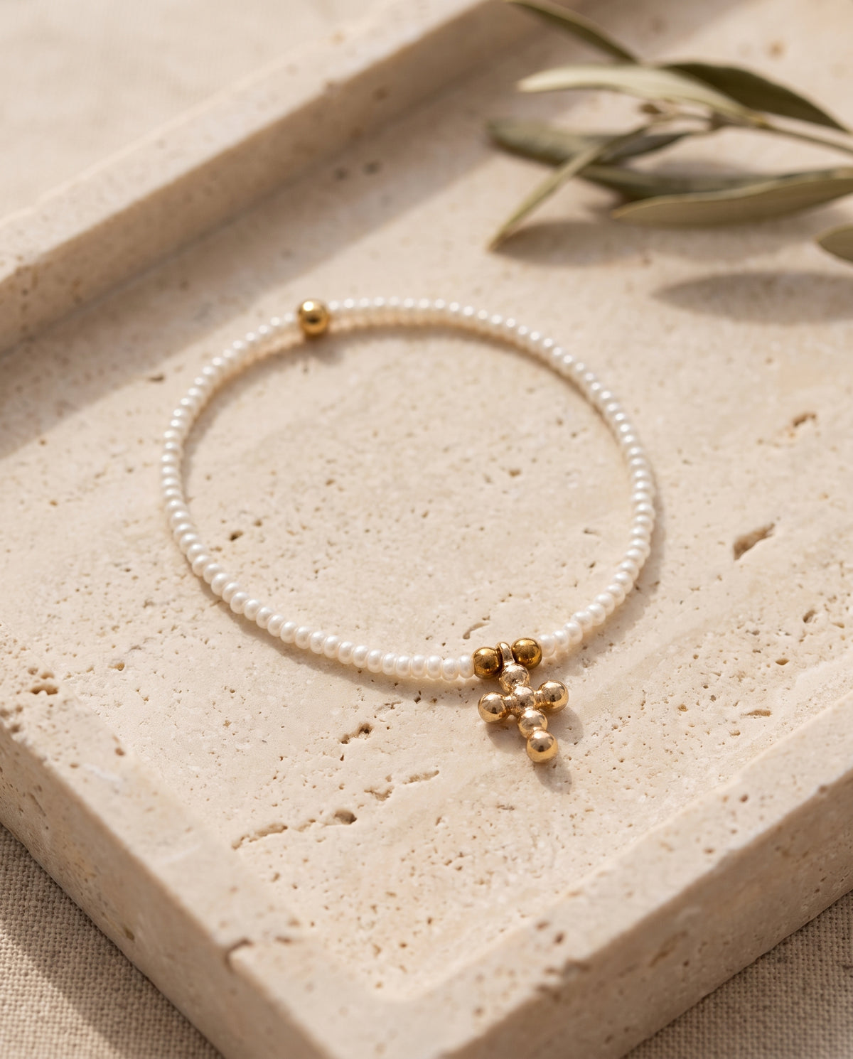Pulsera cruz de perla natural blanca sobre bandeja de piedra beige con rama de olivo, Perla Santa