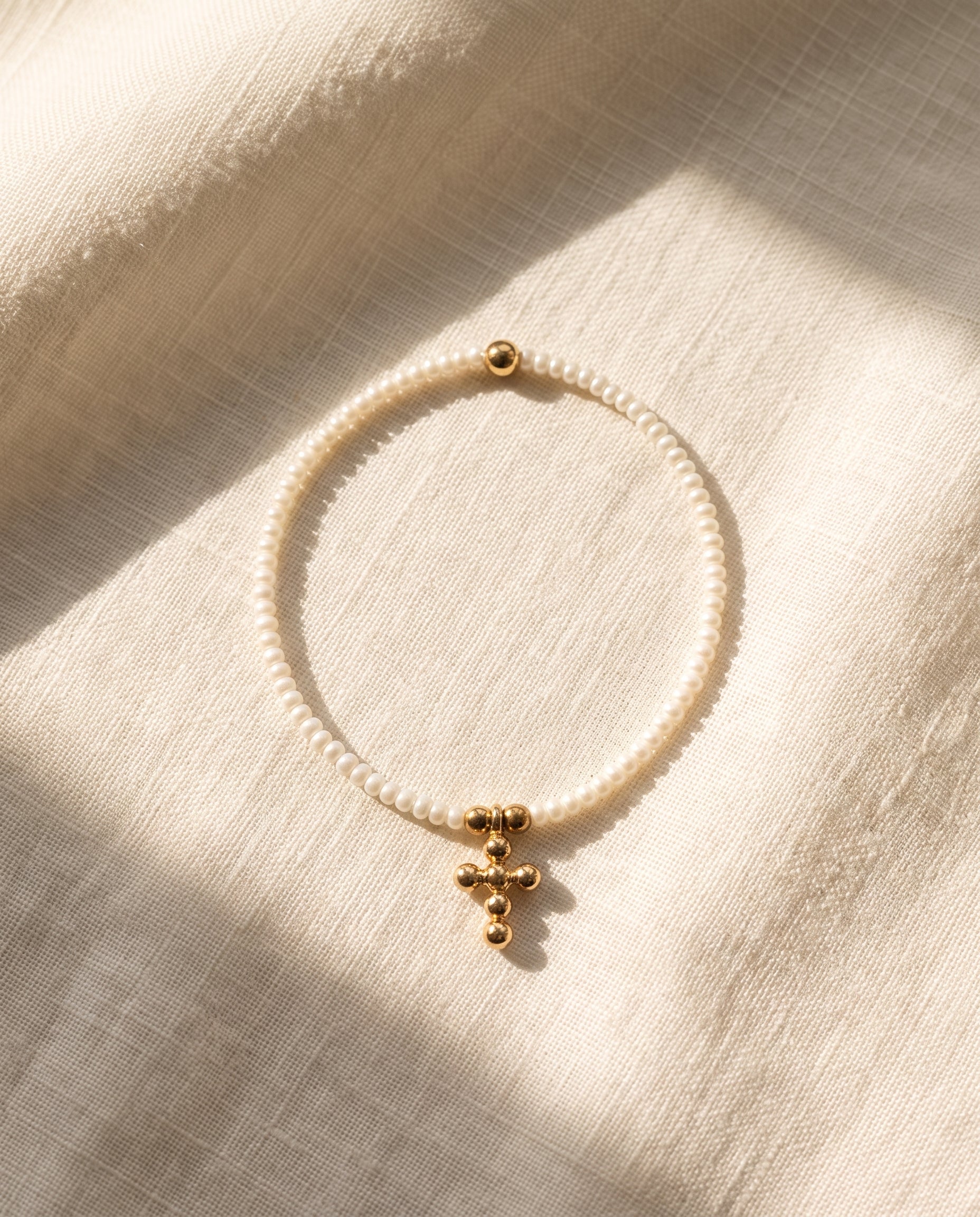 Pulsera cruz de perla natural blanca sobre lino crema con luz calida, Perla Santa