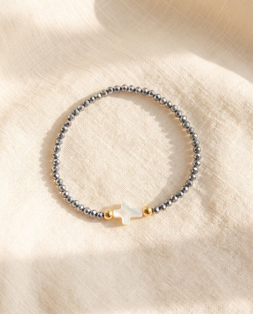 Pulsera con cruz de concha nacar sobre lino crema con luz calida, Perla Santa
