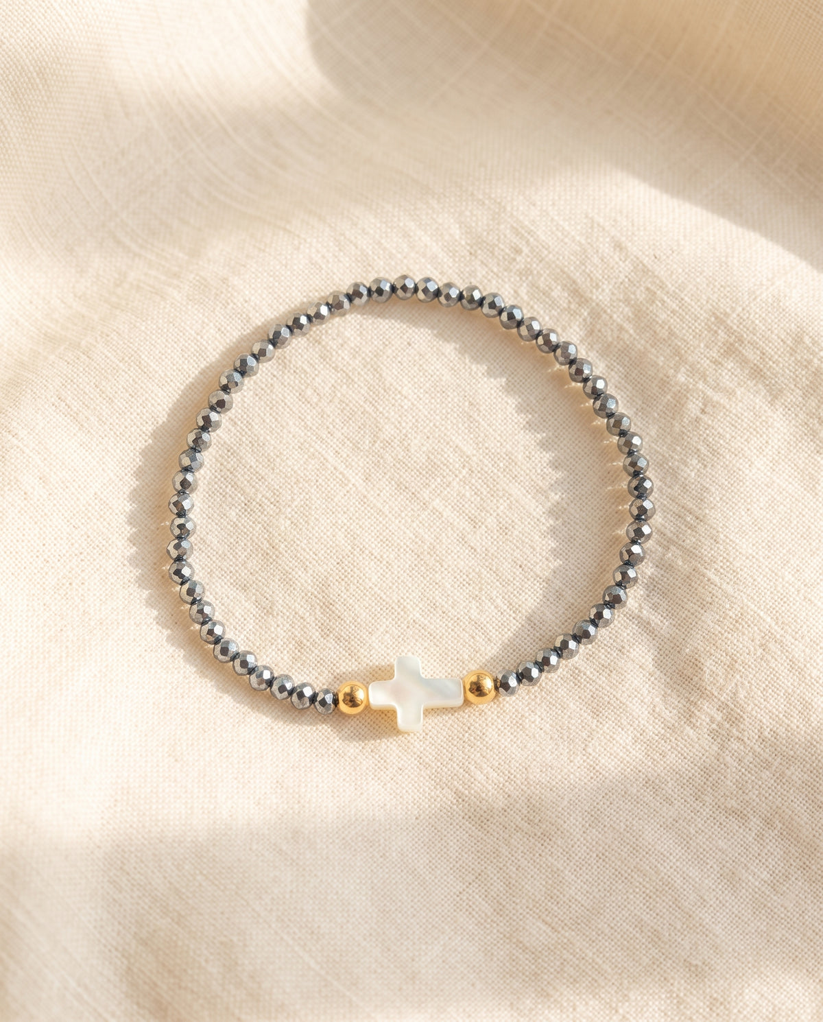 Pulsera con cruz de concha nacar sobre lino crema con luz calida, Perla Santa