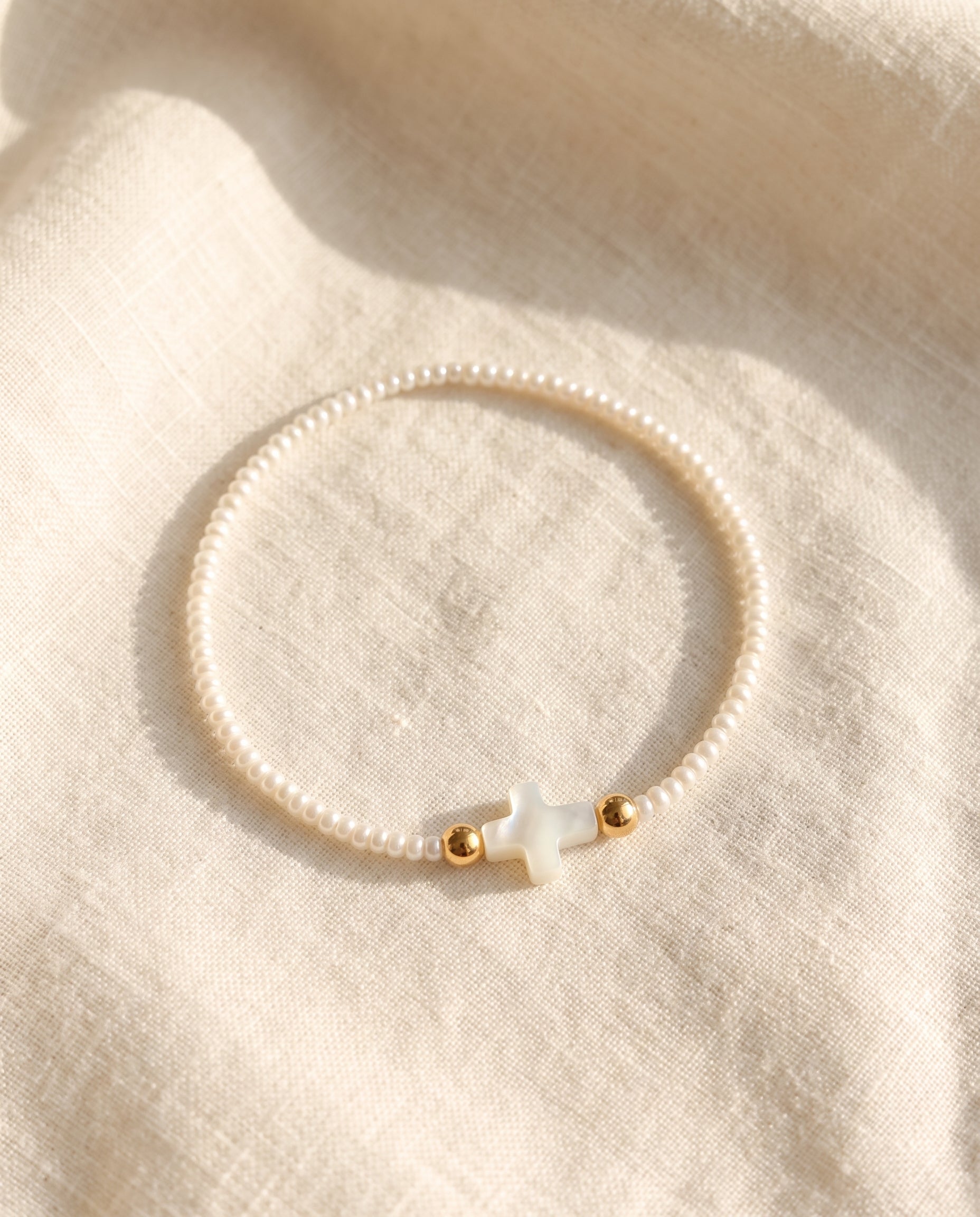 Pulsera de perla con cruz de concha nacar sobre lino crema con luz calida, Perla Santa